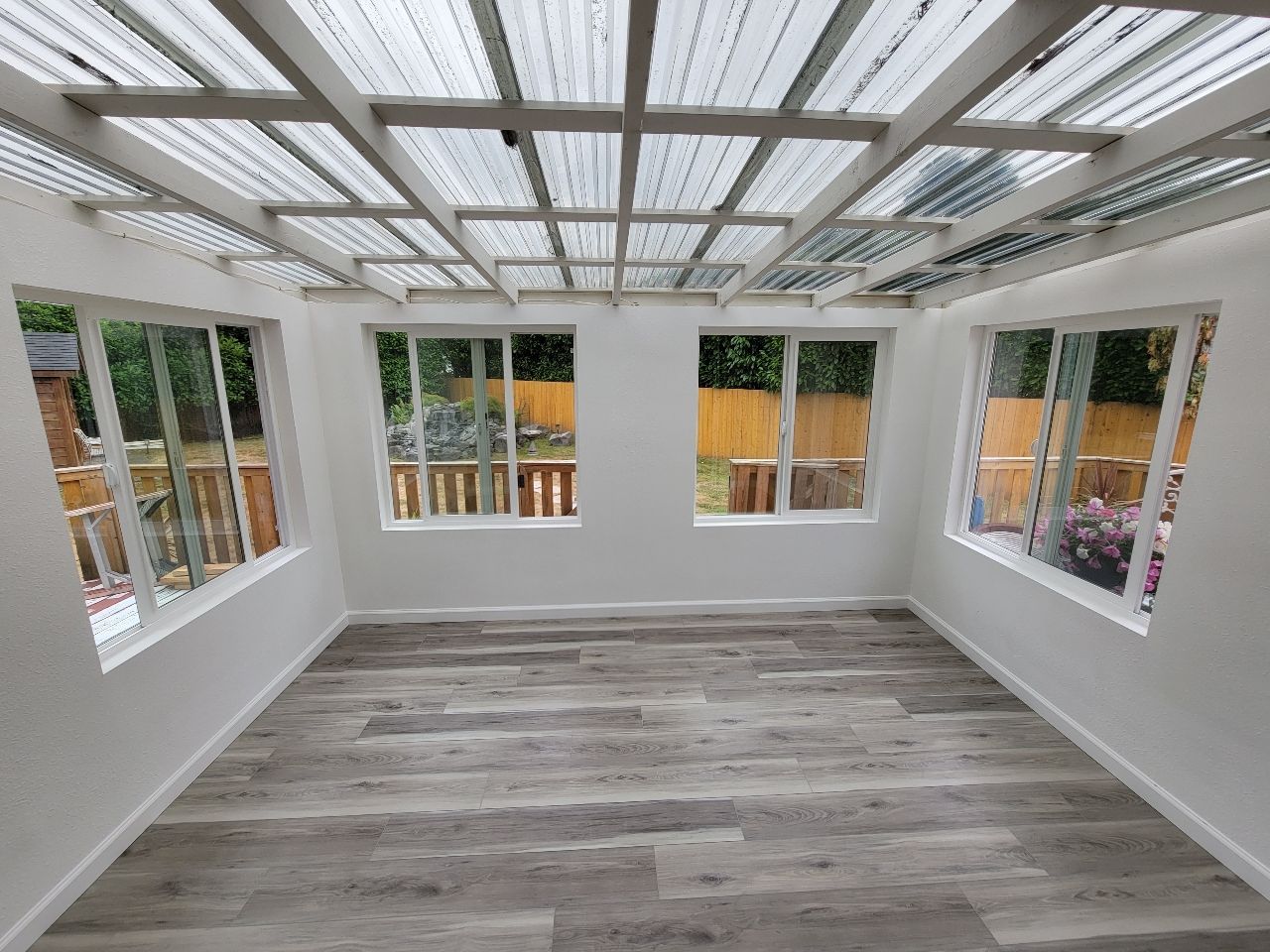 Empty sunroom with white walls, windows, and a corrugated plastic ceiling. Gray wood-look flooring.