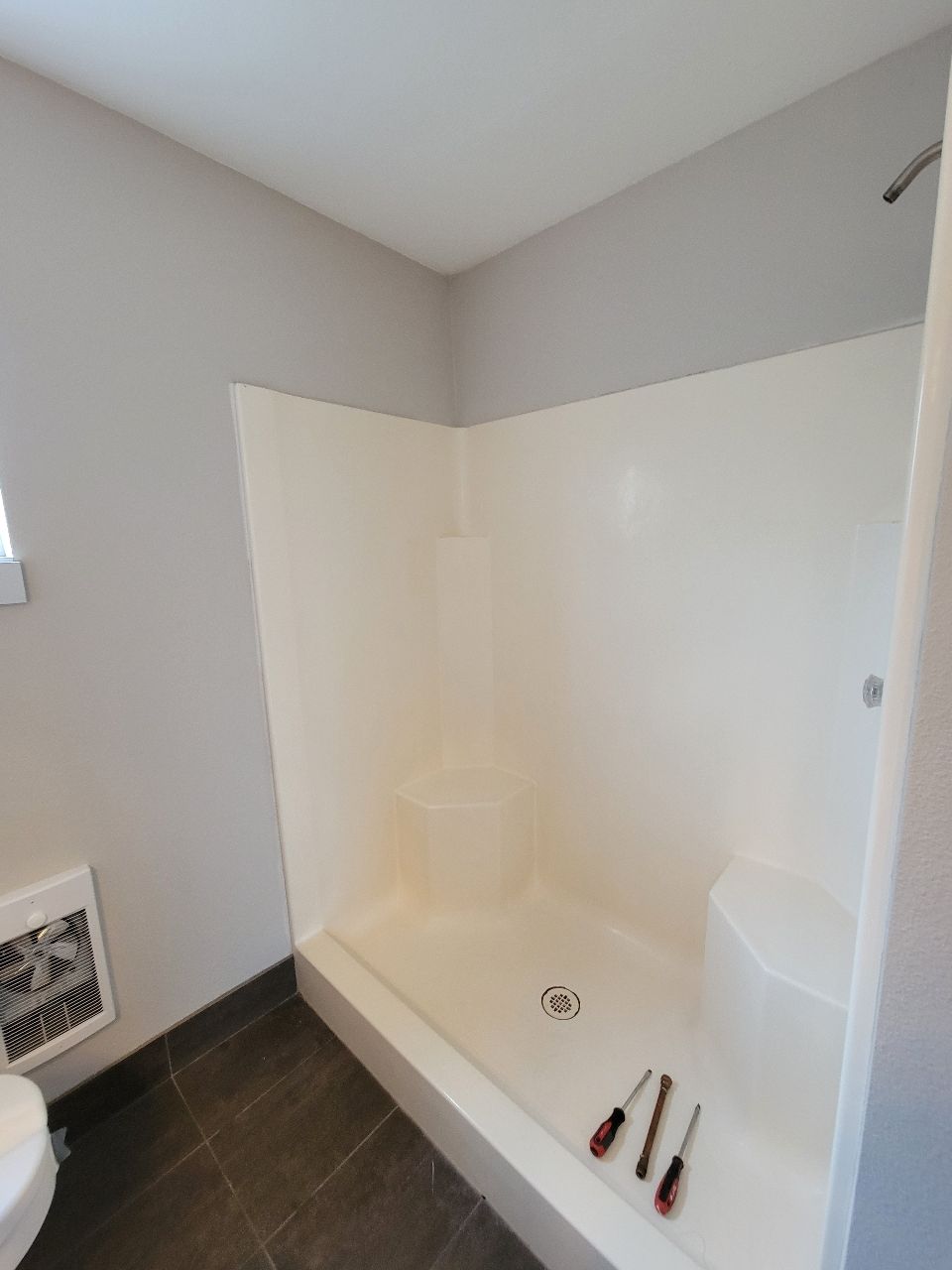 Empty, corner shower stall in a bathroom with grey walls, dark tile floor. Two screwdrivers on floor.