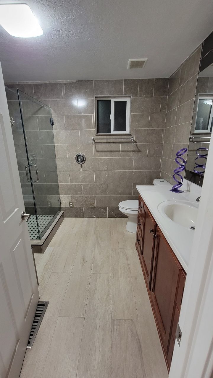 Bathroom with tile walls and floor, shower, toilet, double vanity, and small window.