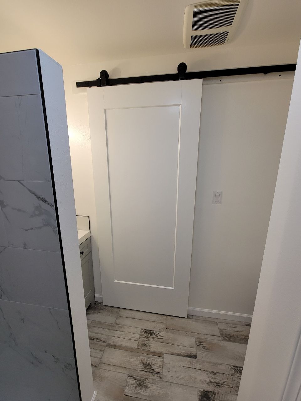 White barn door in a bathroom, set on a black track, against white walls and a wood-look floor.