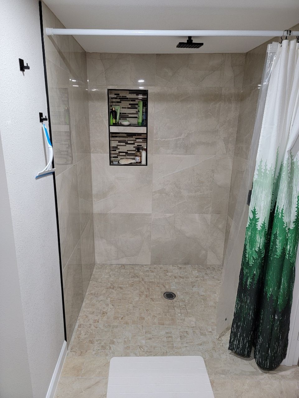 Shower stall with beige tiles and glass door. Green curtain on the right. White mat on the floor.