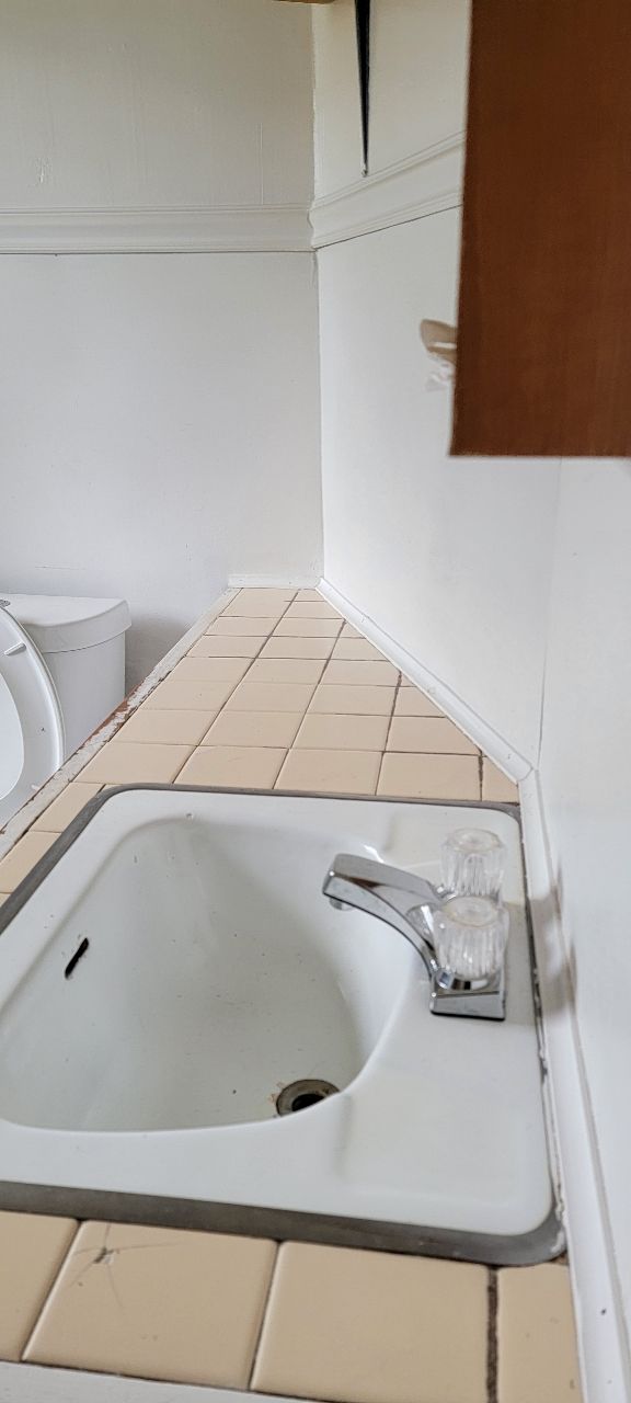 A bathroom sink with a tiled countertop. White walls, a chrome faucet, and a small brown cabinet.