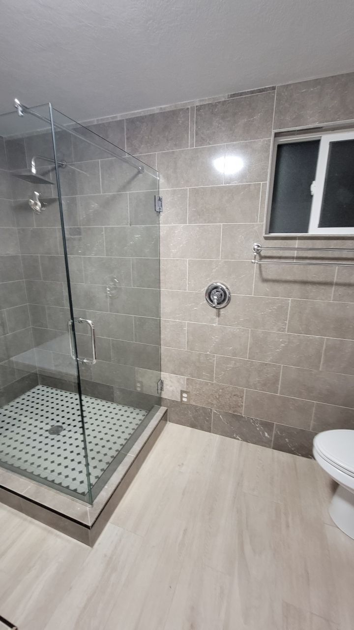 A remodeled bathroom with a glass shower, gray tile, and a toilet. The floors are a light tan color.