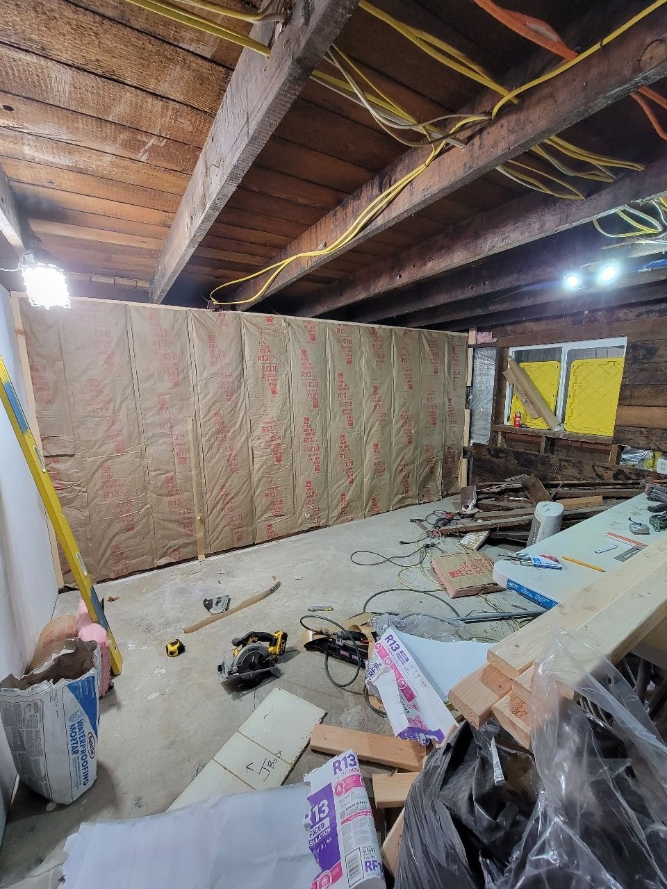 Interior construction view: a wall with insulation installed, tools, and debris on the concrete floor.