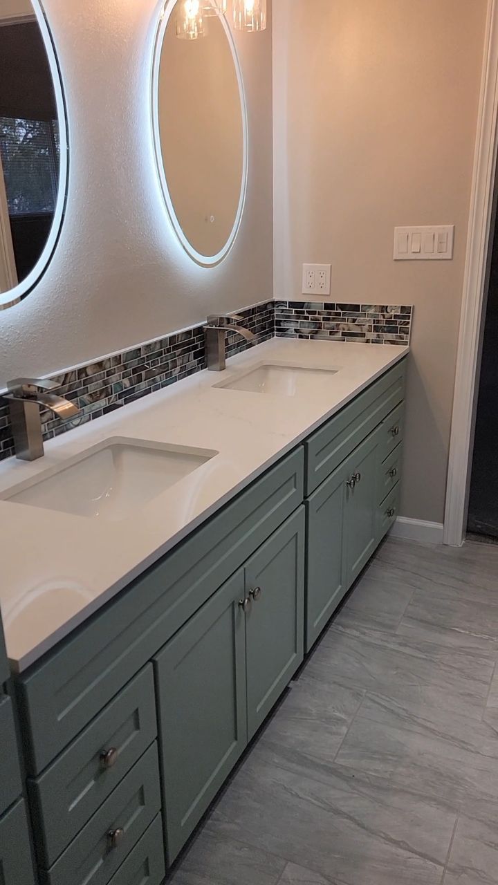 Bathroom with blue-green cabinets, white countertop, mosaic backsplash, two sinks, and illuminated mirrors.