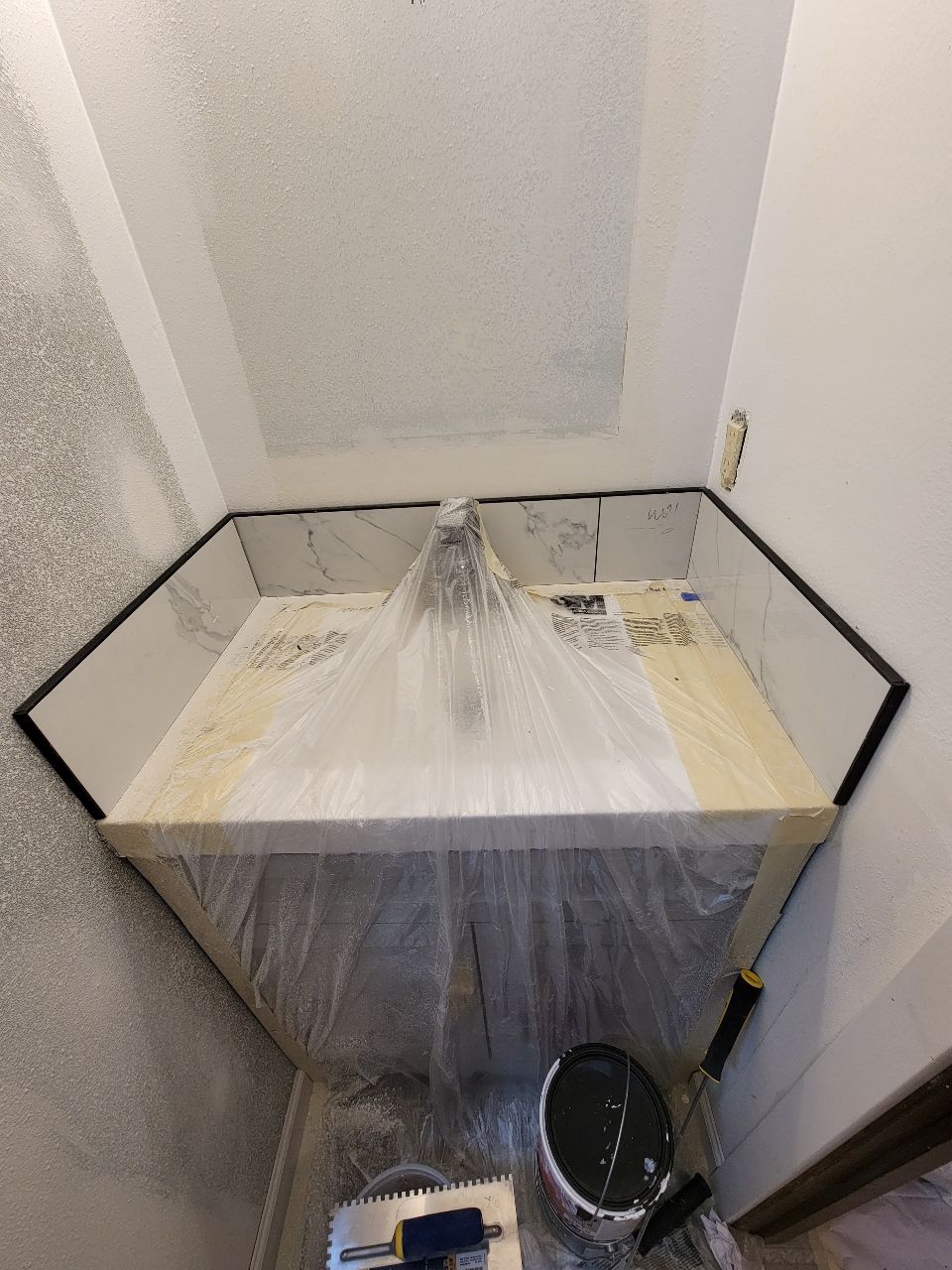 A newly tiled shower stall, hexagonal shape. Marble-look tiles. Plastic sheeting covers the floor. Black trim.