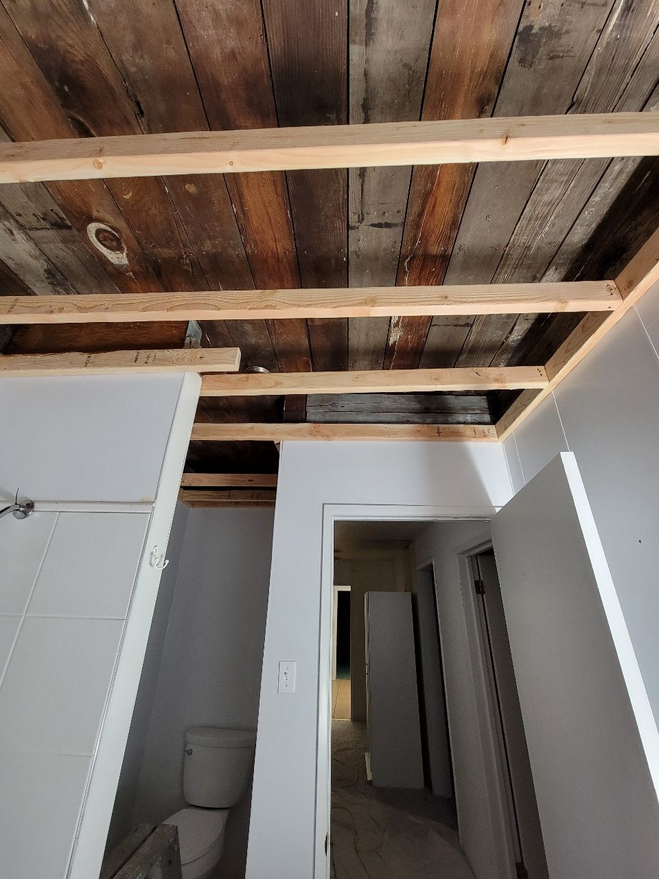 Bathroom under renovation. Wood plank ceiling with exposed support beams. White walls and doorway leading to hallway.