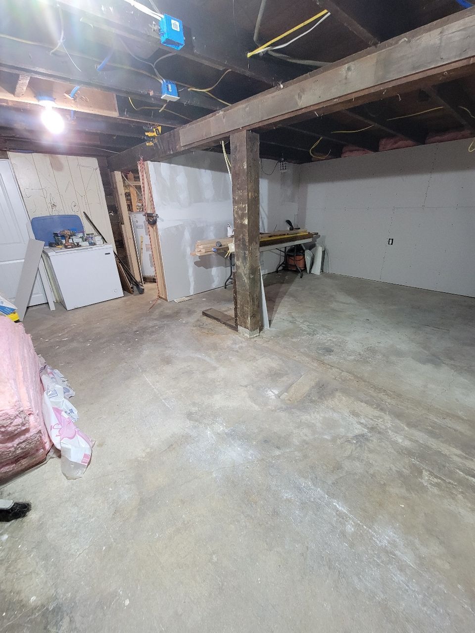 Basement with exposed beams, concrete floor, drywall, and support post. Tools and materials are scattered.