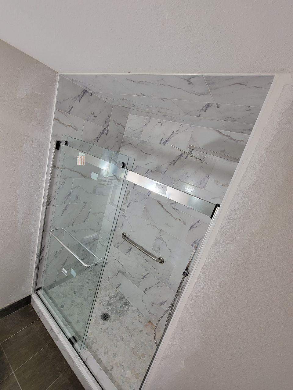 Marble-tiled shower with glass doors and a chrome handle, in a corner space with gray walls.