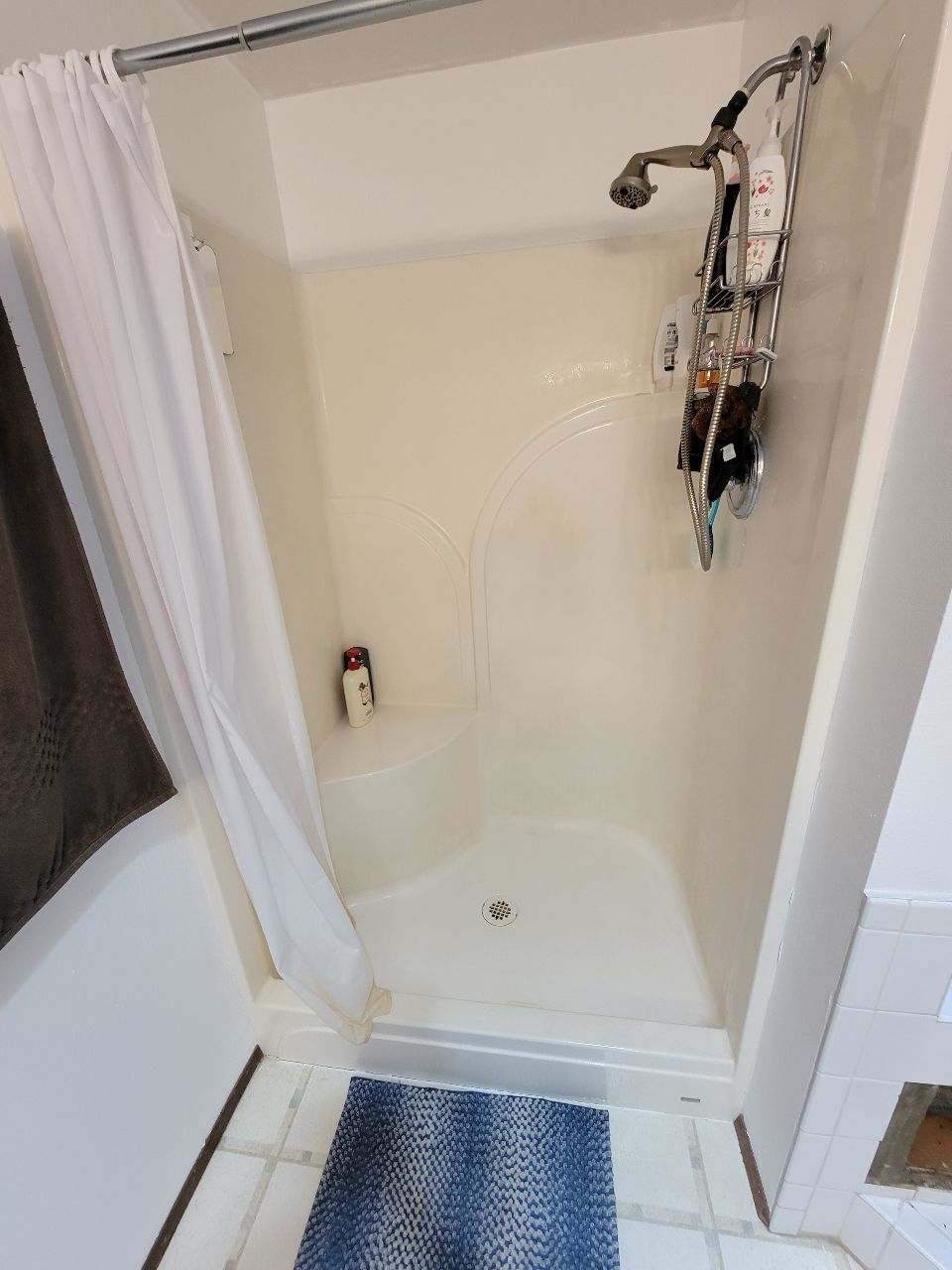 Cream-colored shower stall with a curtain, a blue rug, and toiletries.