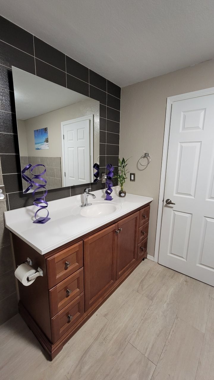 Bathroom with wood vanity, white countertop, large mirror, dark tile, and white door.