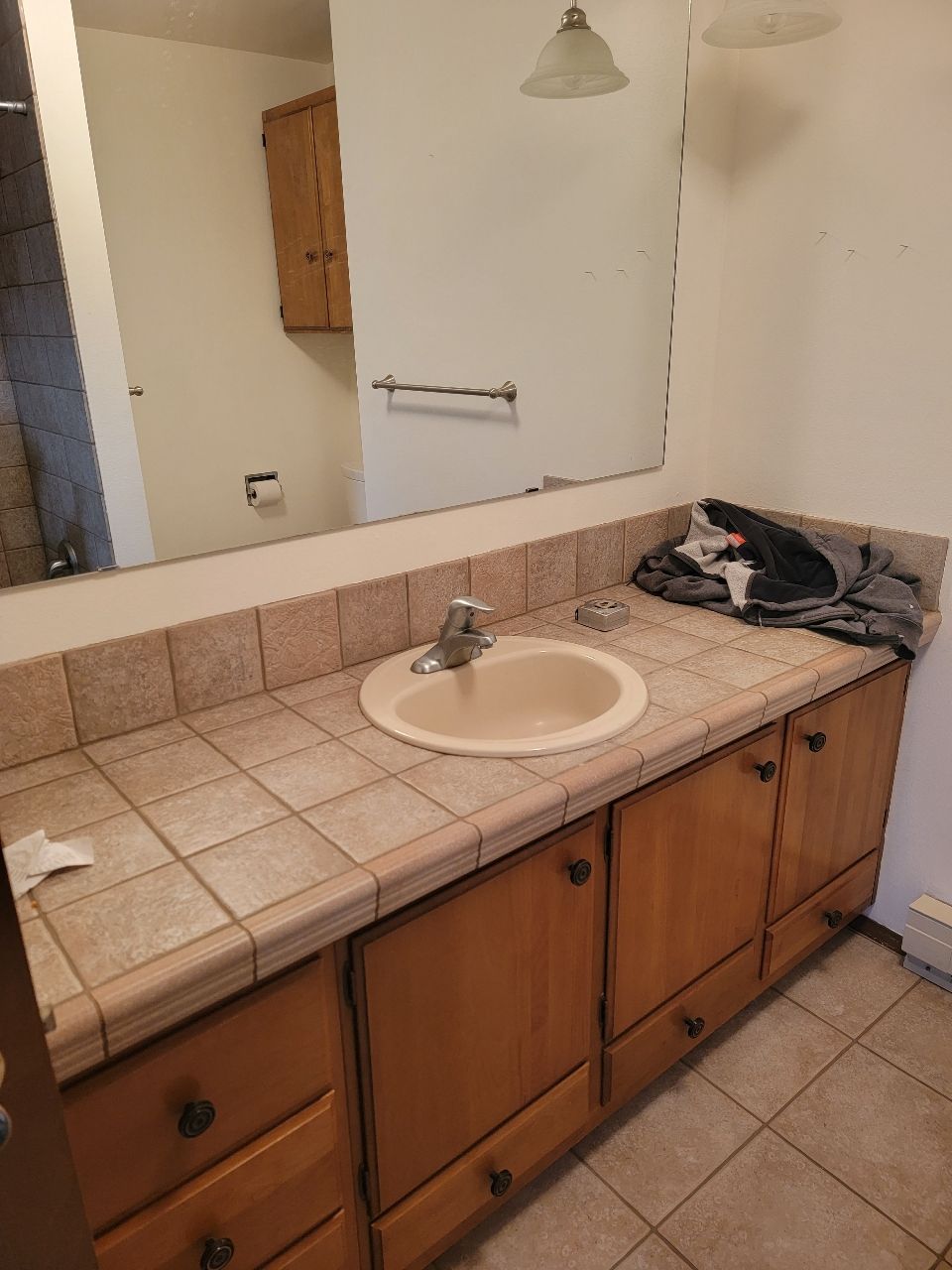 Bathroom with beige tiled countertop, wooden cabinets, oval sink, mirror, and towel bar.