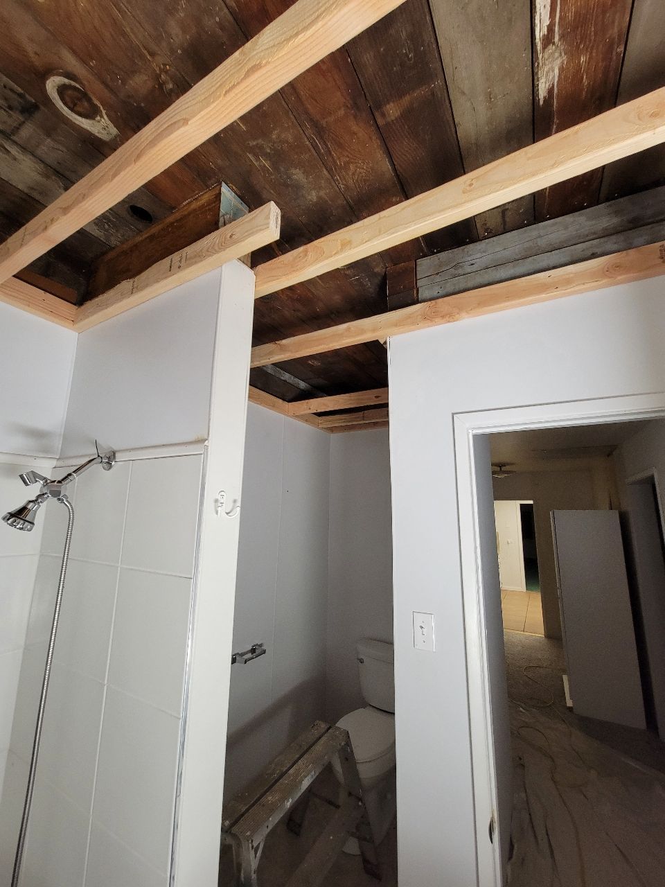 Bathroom renovation in progress, showing exposed wood ceiling beams above a toilet, white walls, and a shower fixture.