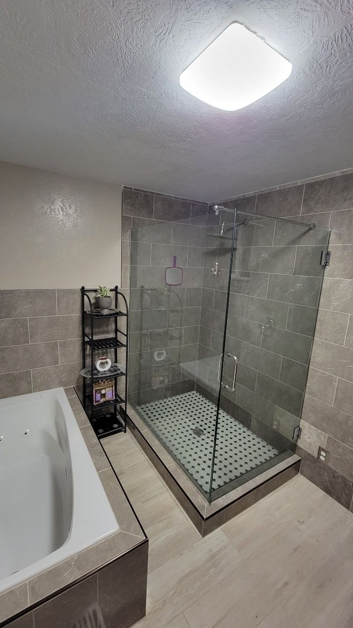 Modern bathroom with gray tile, glass shower, white tub, and black shelving.