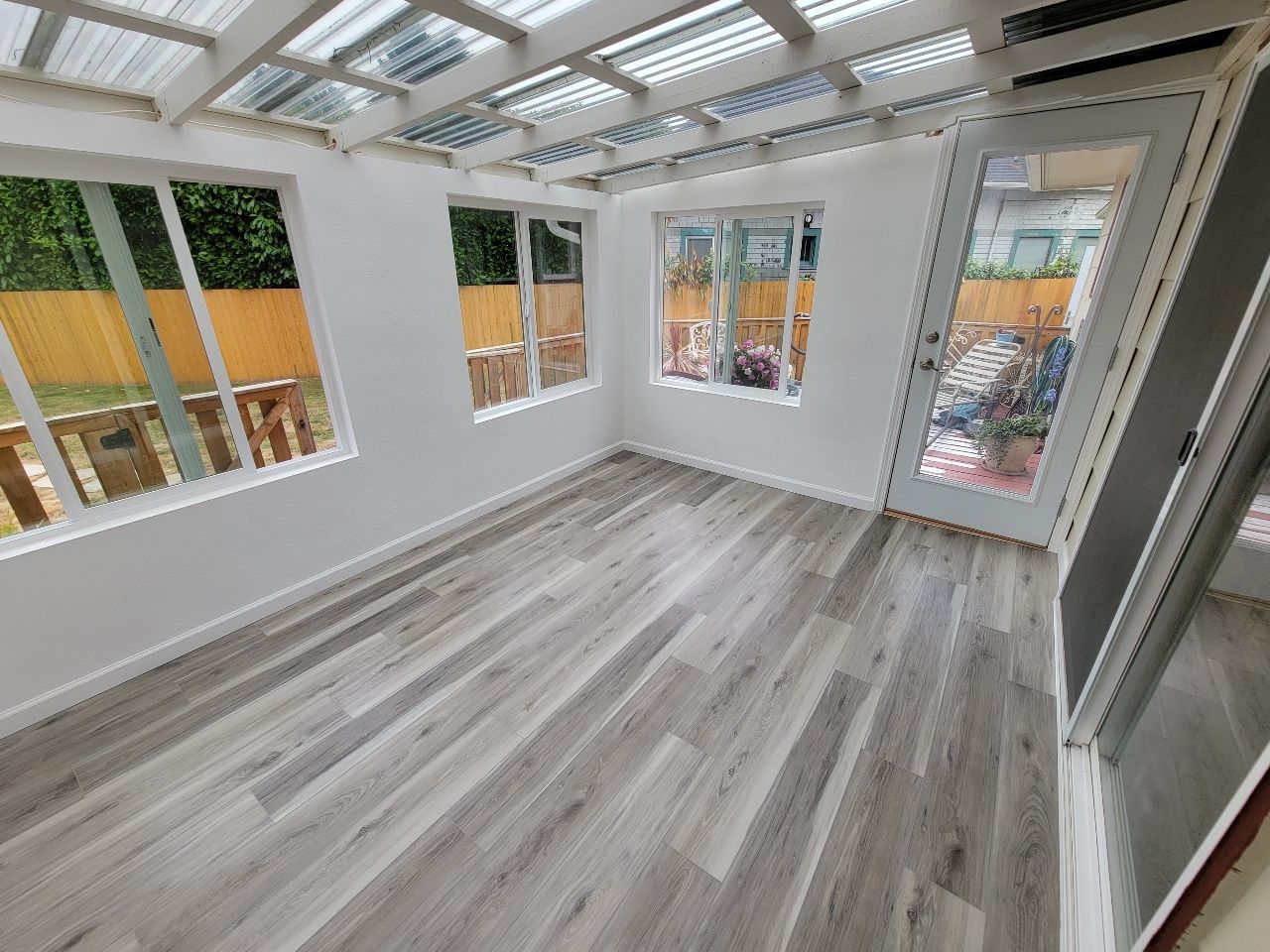 Sunroom with gray wood-look floors, white walls, and clear roof panels, windows showing a deck and door to the outdoors.