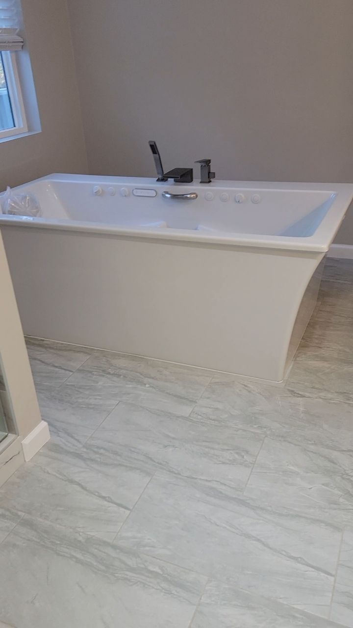 White freestanding bathtub in a light-colored tiled bathroom. Faucet at top, window in background.