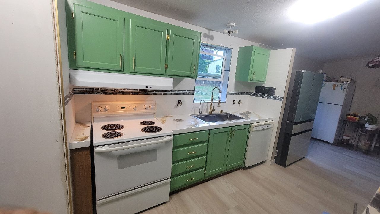 Green-painted kitchen cabinets and white appliances, including a stove and refrigerator, sit in a kitchen with light-colored flooring.
