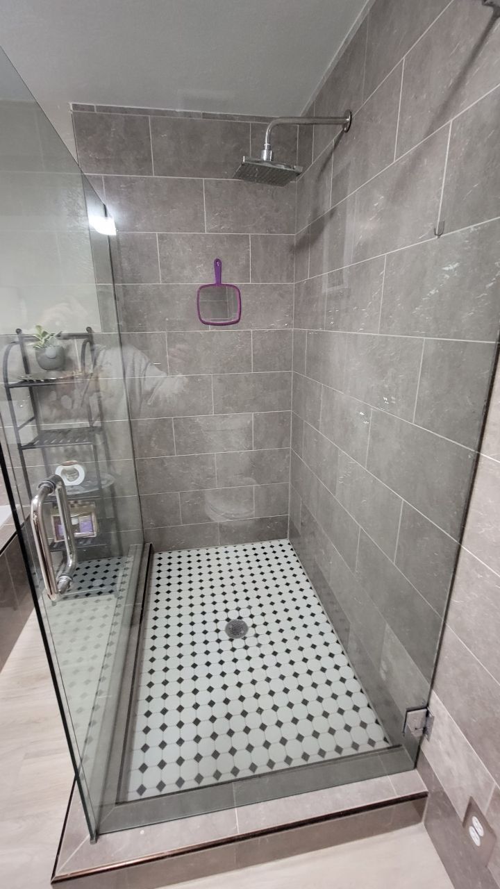 Walk-in shower with gray tile walls, glass door, and a checkered floor. A shelf and showerhead are visible.