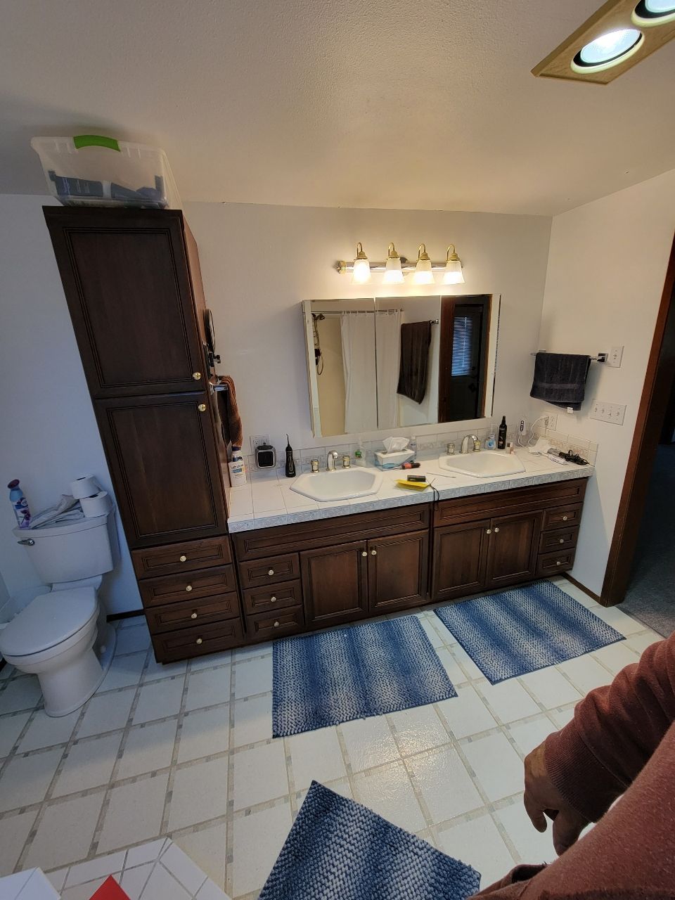 Bathroom with a double sink vanity, tall storage cabinet, and toilet. Blue rugs and white tile floor.