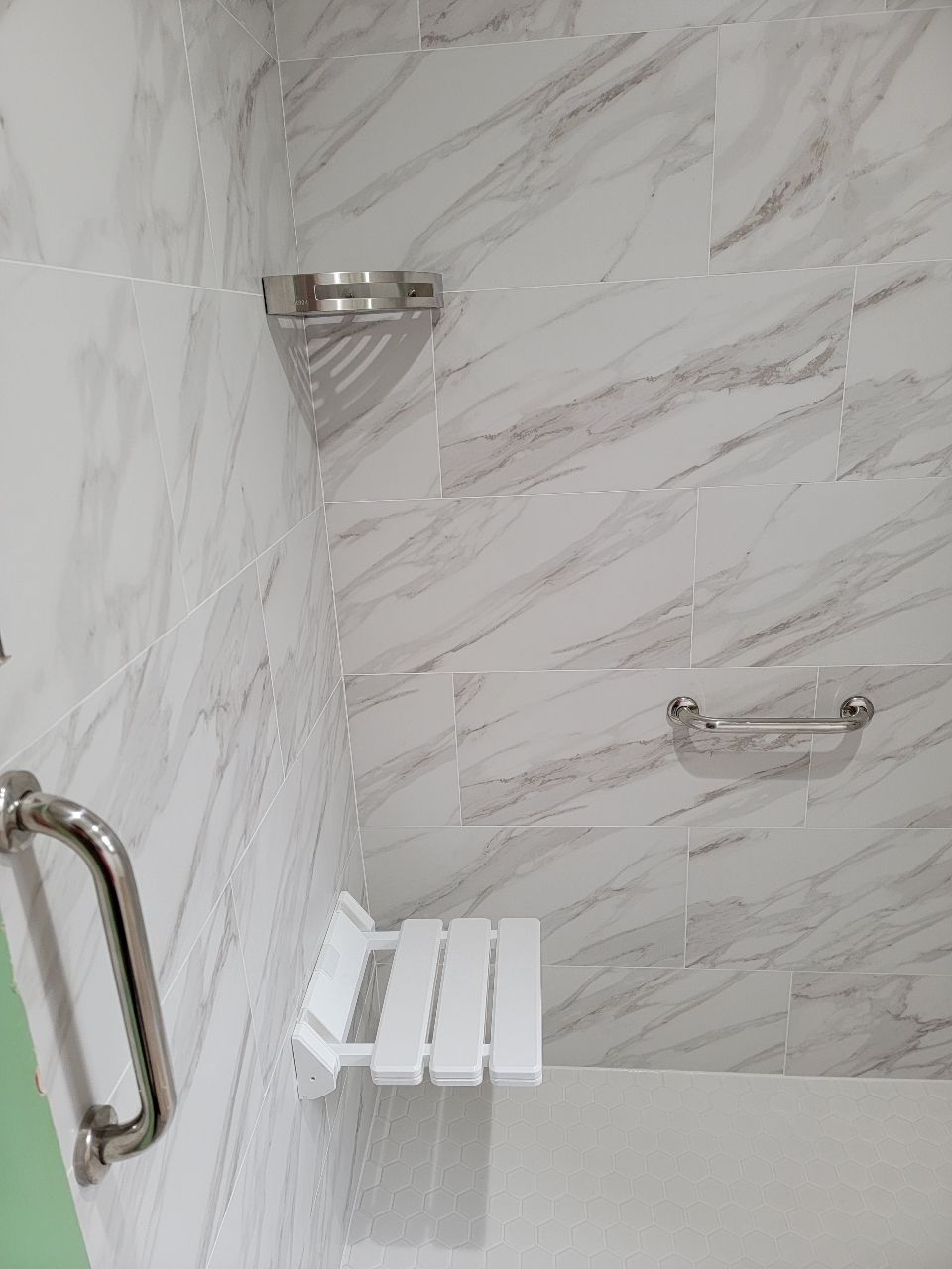 Shower with white marble-like tile, a corner shelf, a grab bar, and a fold-down seat.