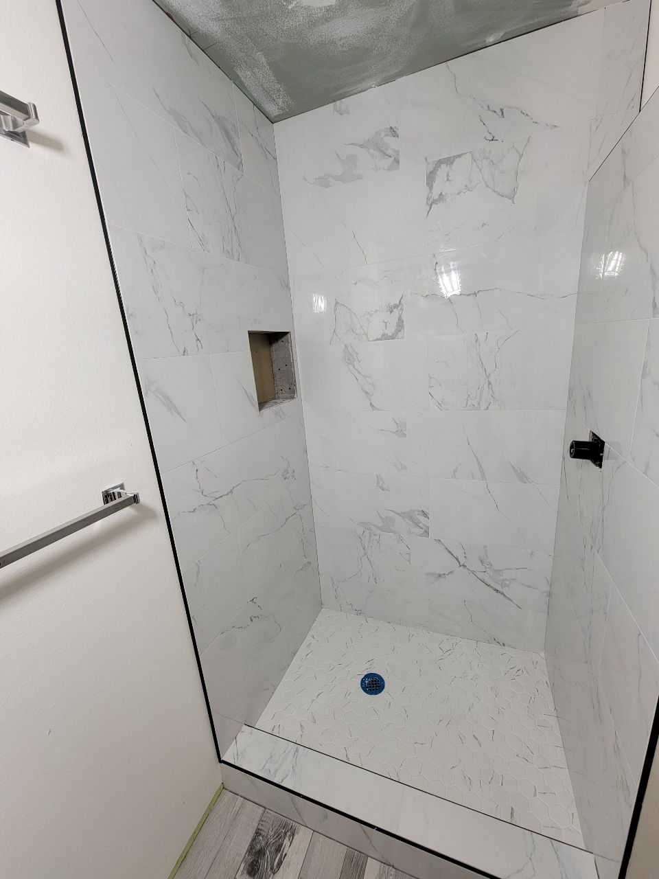 White tiled shower with marble-like veining, black trim, niche, and blue drain.