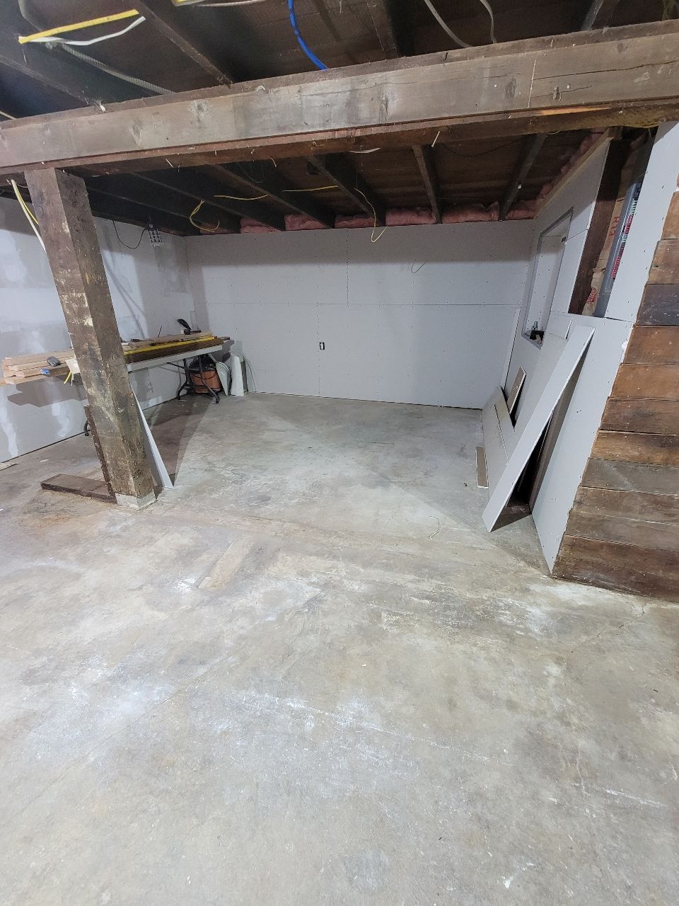 Basement renovation in progress, concrete floor, white walls, exposed beams, lumber leaning against wall.