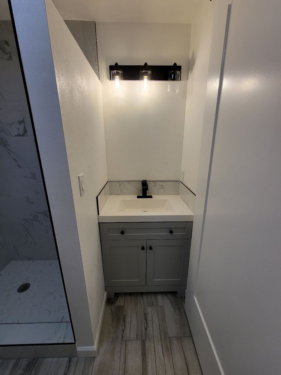 Narrow bathroom with gray vanity, white countertop, black faucet, and three-bulb light fixture.