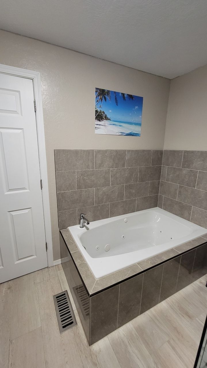 Bathroom with a white bathtub surrounded by gray tiles, a beach scene picture, and light wood-look flooring.