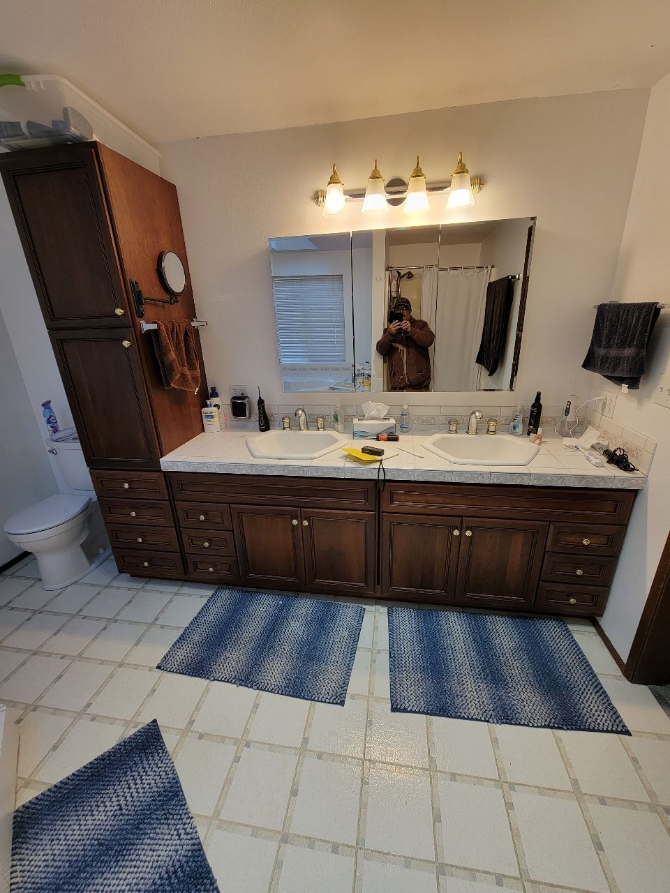 Bathroom with a double vanity, dark wood cabinets, and blue rugs. A tall cabinet and toilet are visible.