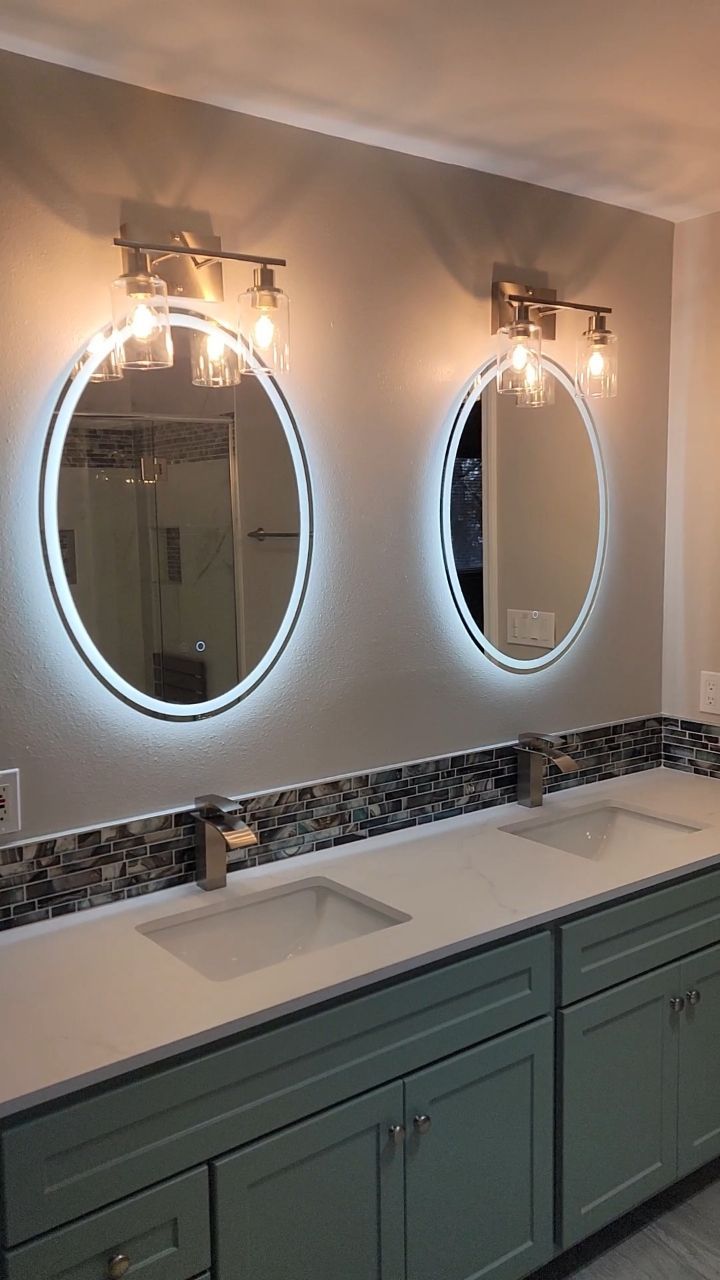 Two illuminated oval mirrors above green vanity with sinks and brown tile backsplash.