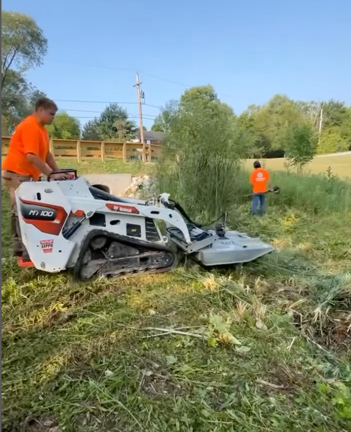 Man operating Bobcat mower to clear overgrown vegetation near a bridge. Another man using a weed whacker.