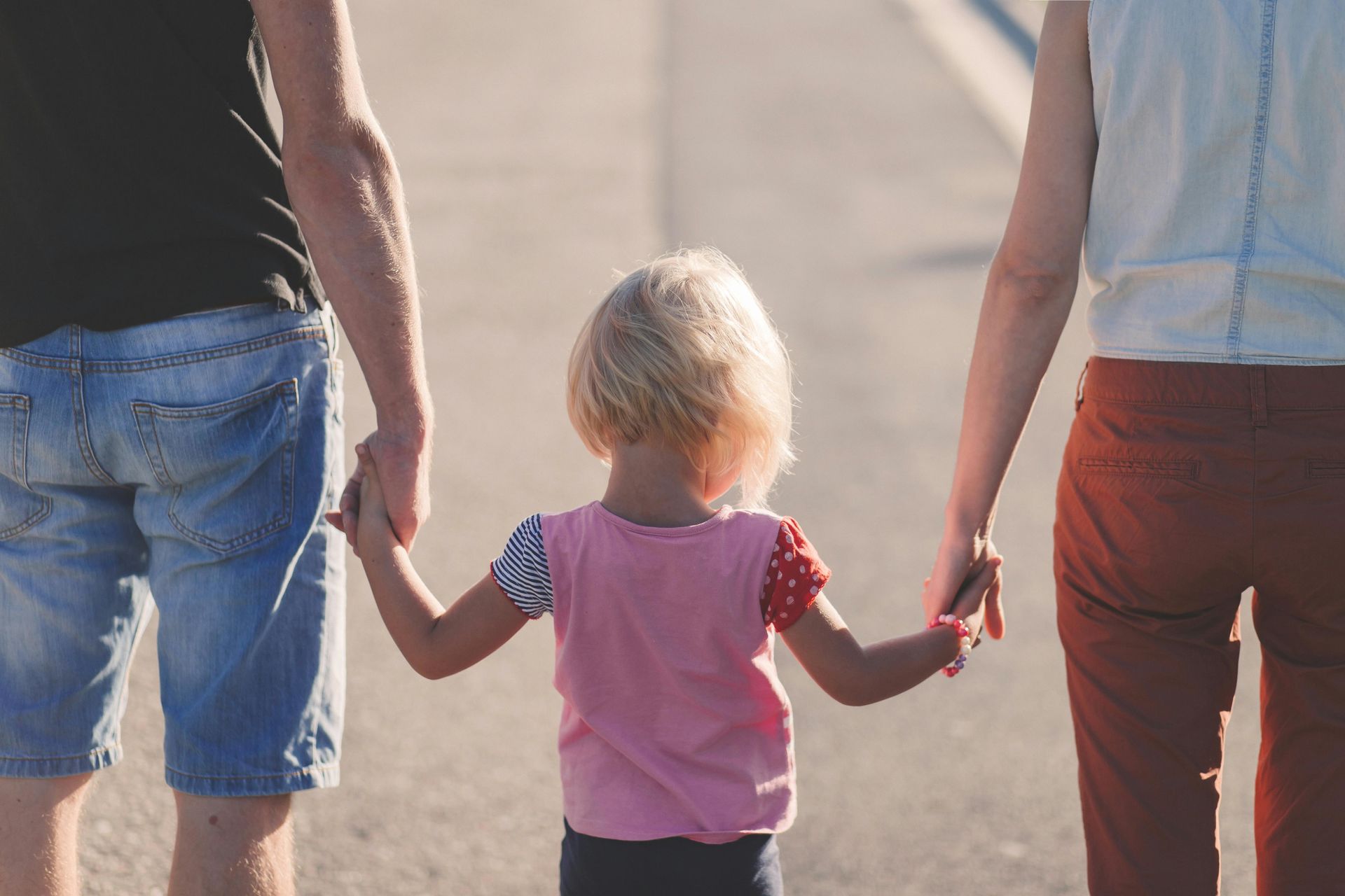 Three people walking hand in hand on a sunlit road, seen from behind.