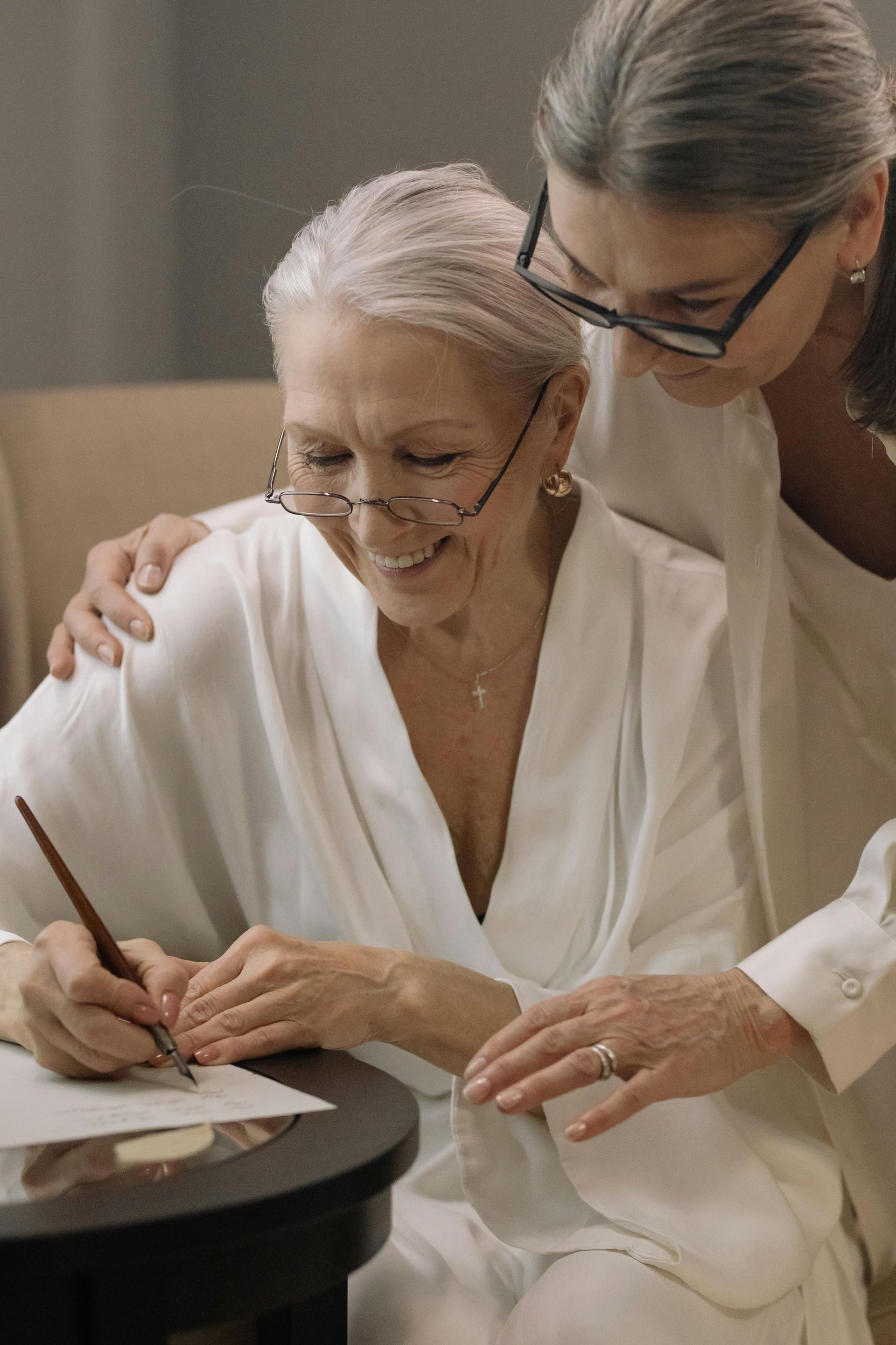 Two women in white robes smiling as one signs a document at a table