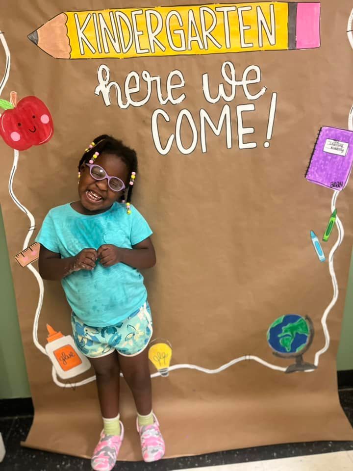A little girl is standing in front of a sign that says kindergarten here we come