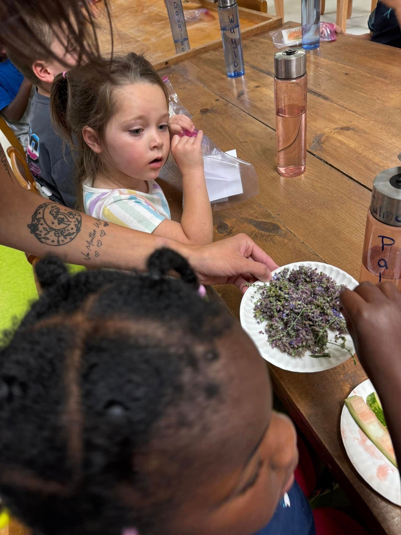 A group of children are sitting at a table looking at a plate of lavender flowers.