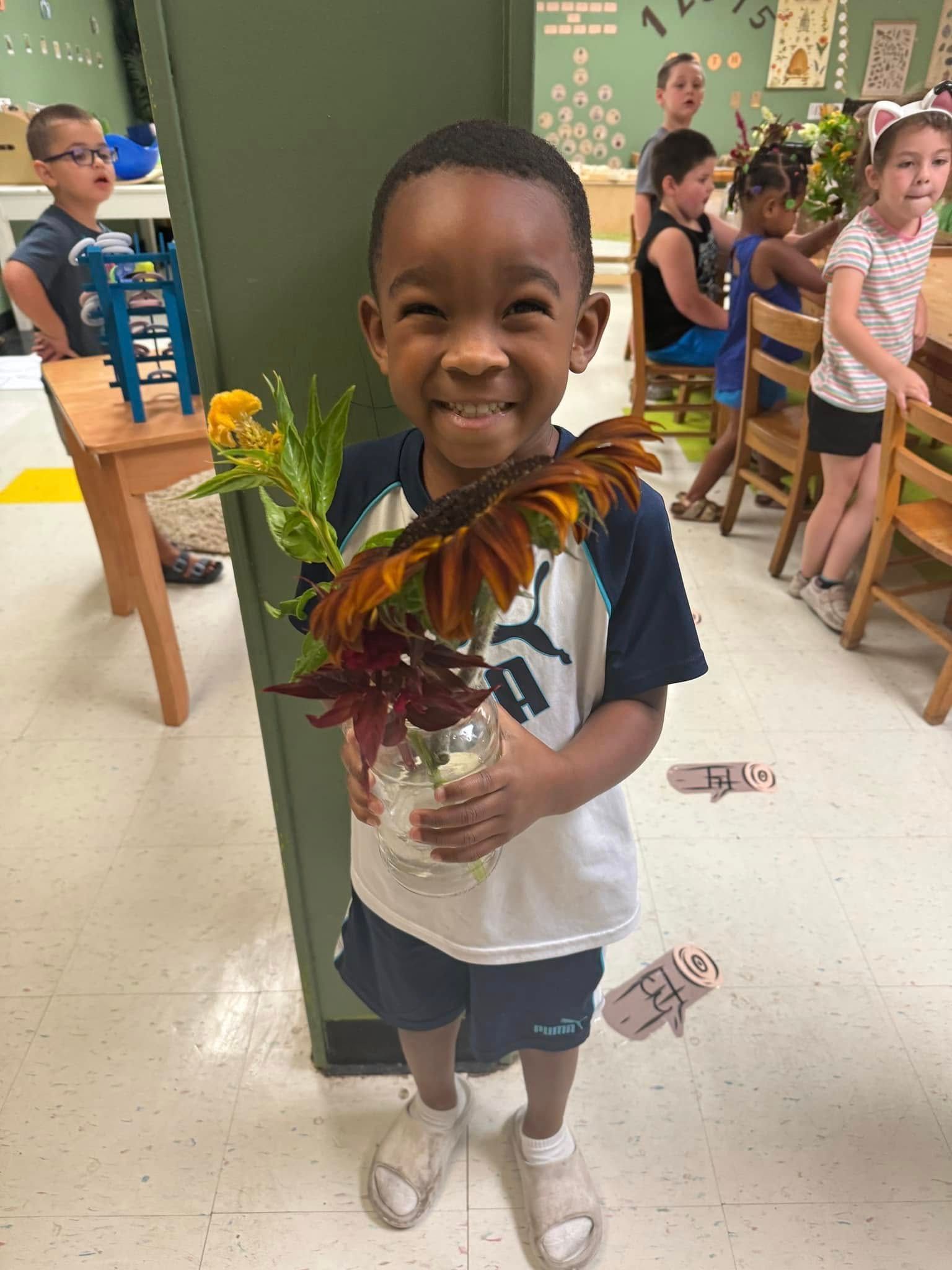 A young boy is holding a vase of flowers in a classroom.