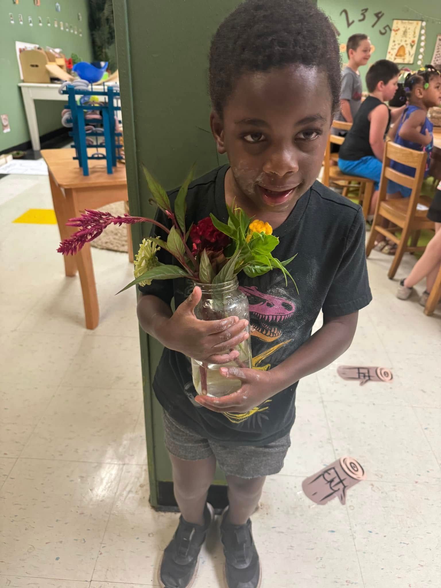 A young boy is holding a vase of flowers in a classroom.