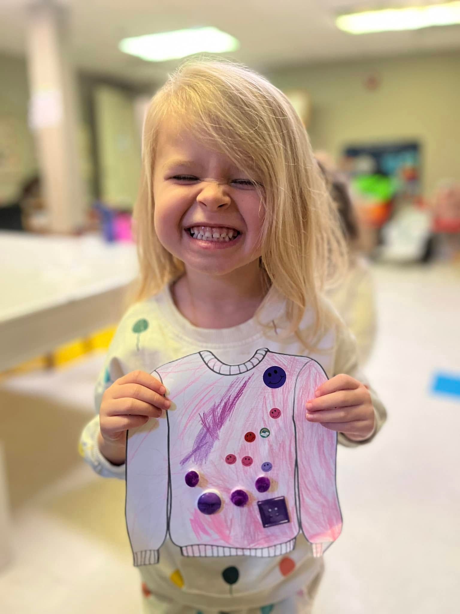 A little girl is holding a drawing of a sweater and smiling.
