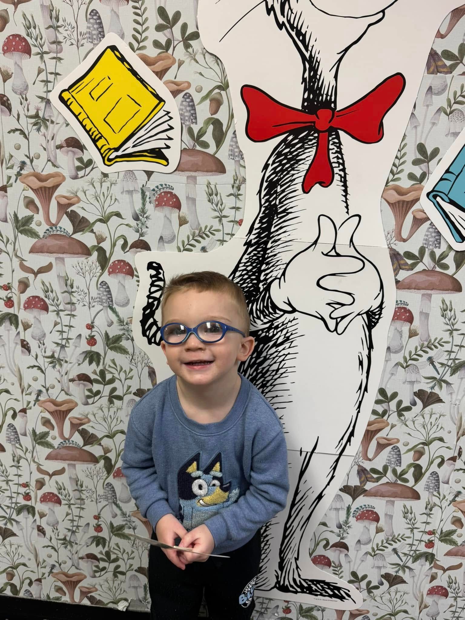 A little boy wearing glasses is standing in front of a cat in the hat statue.