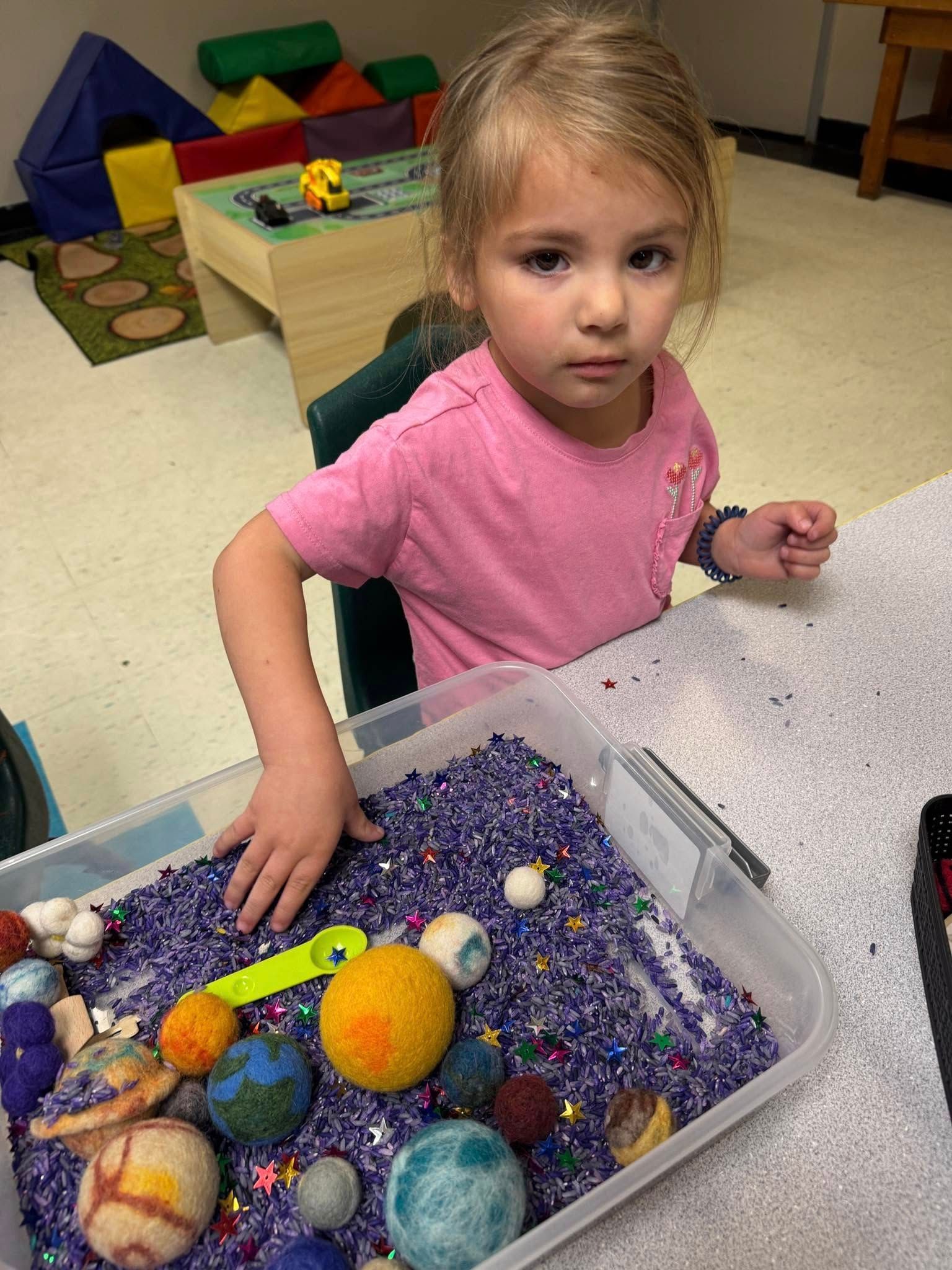 A little girl is sitting at a table playing with a sensory bin filled with purple rice.