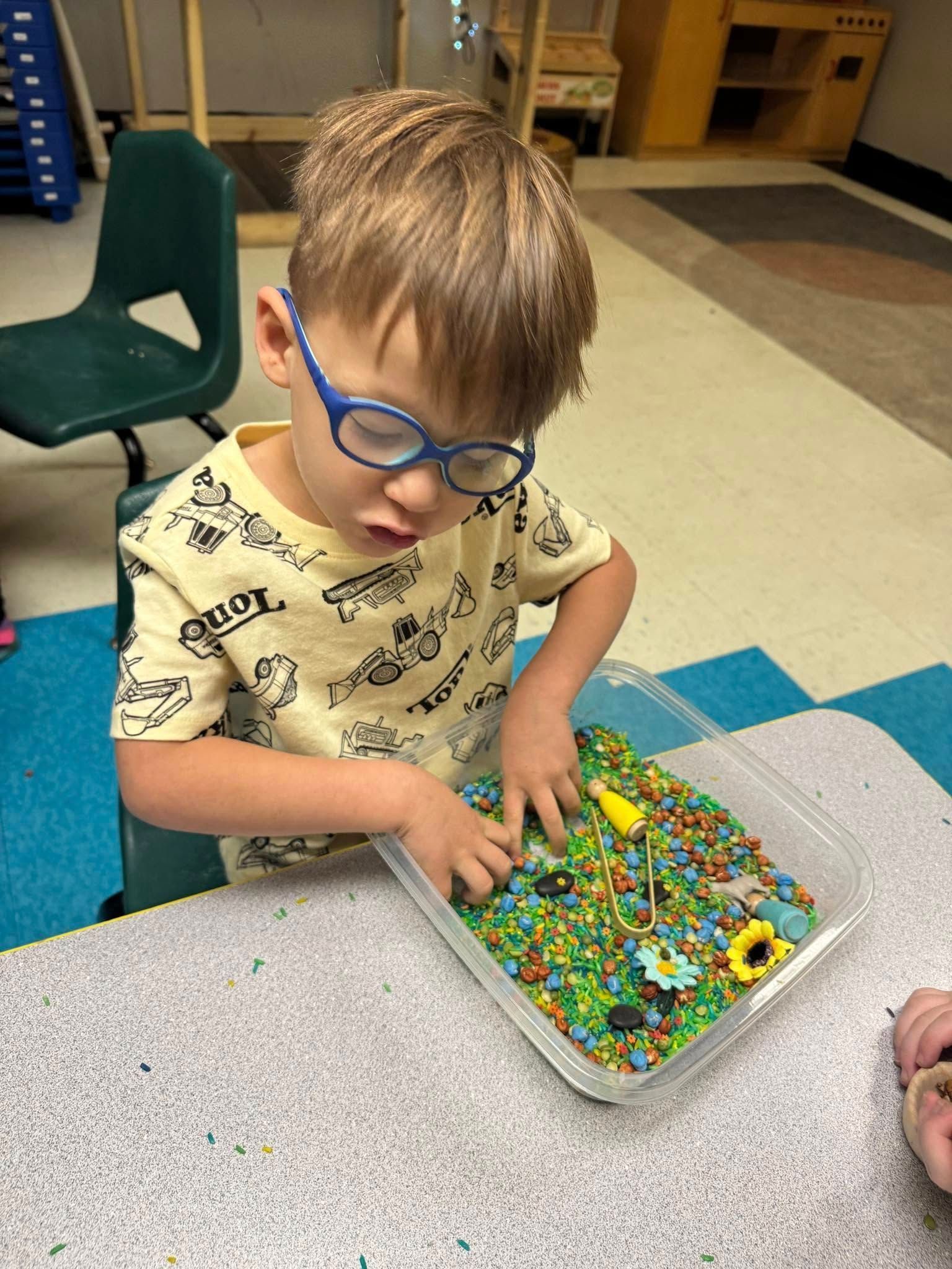 A young boy wearing glasses is playing with a tray of sensory rice.