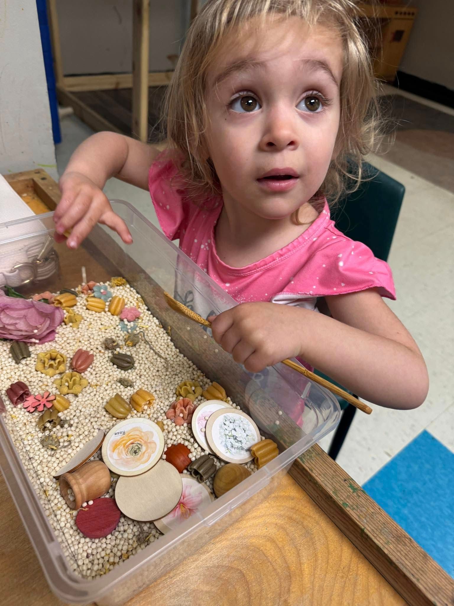 A little girl is sitting at a table playing with a container of rice.