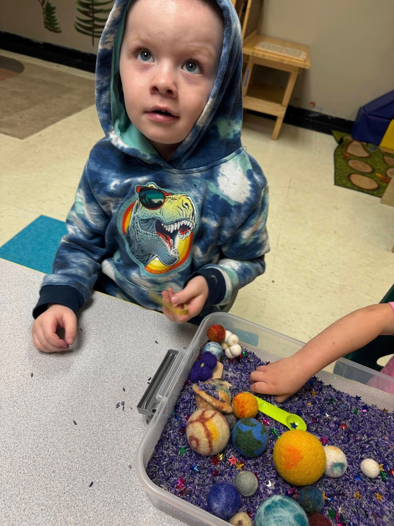 A young boy is sitting at a table playing with a sensory bin filled with purple rice.