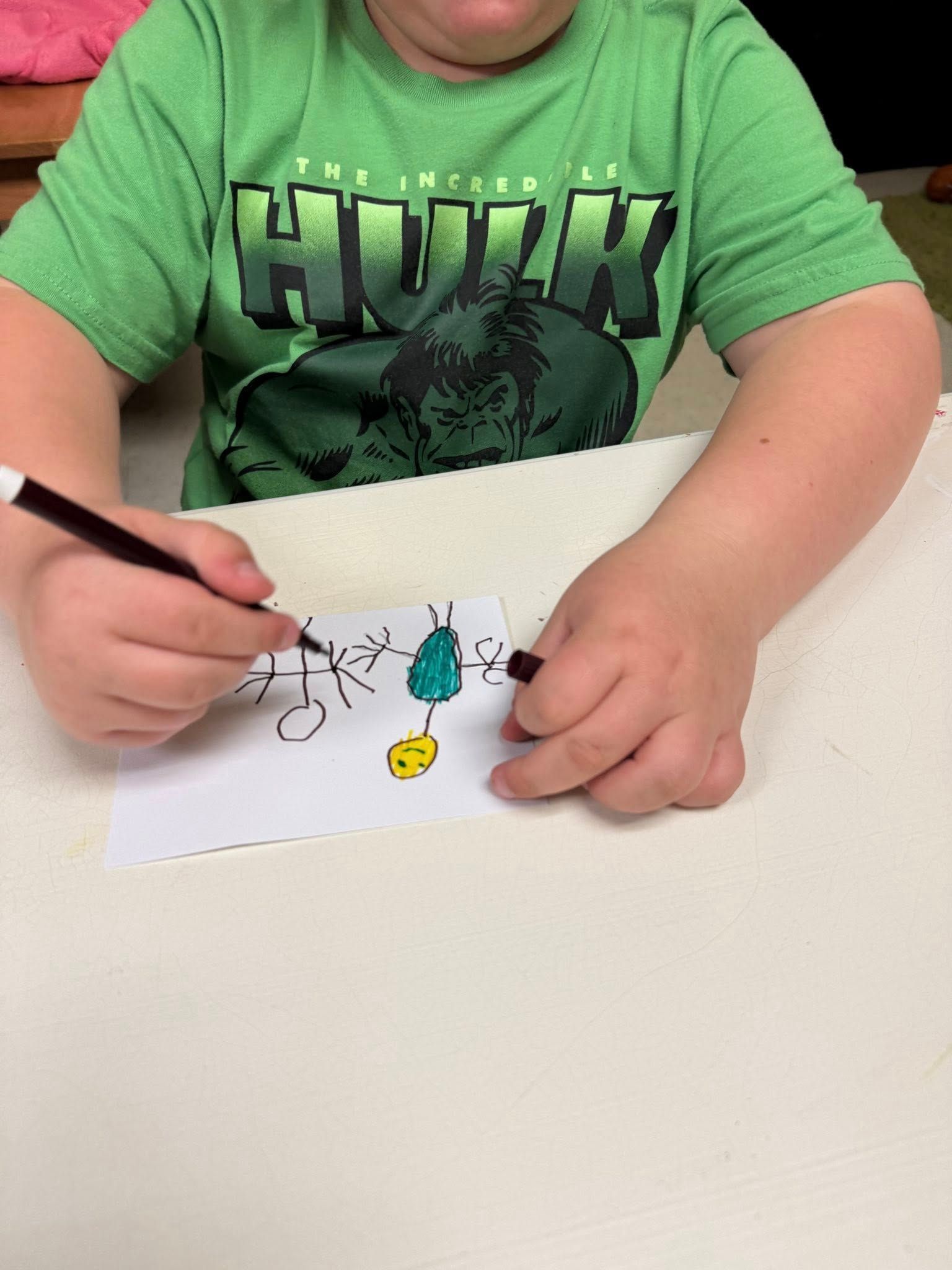 A child wearing a green hulk shirt is drawing on a piece of paper