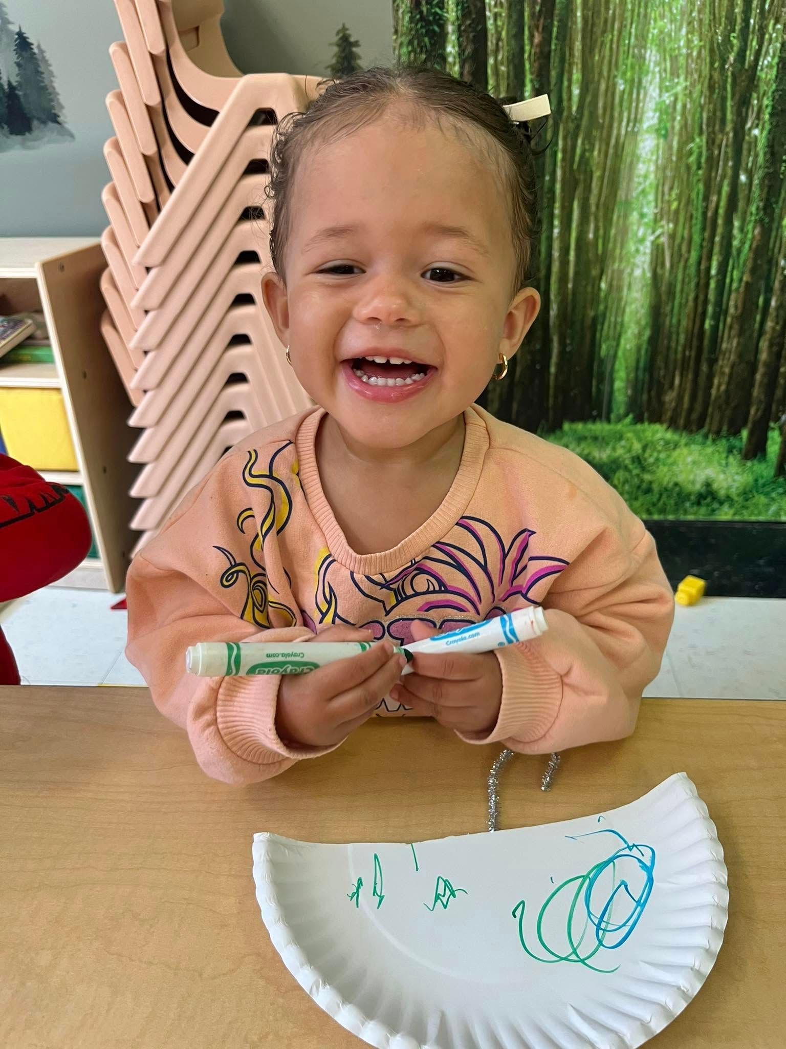A little girl is sitting at a table holding a marker and a paper plate.