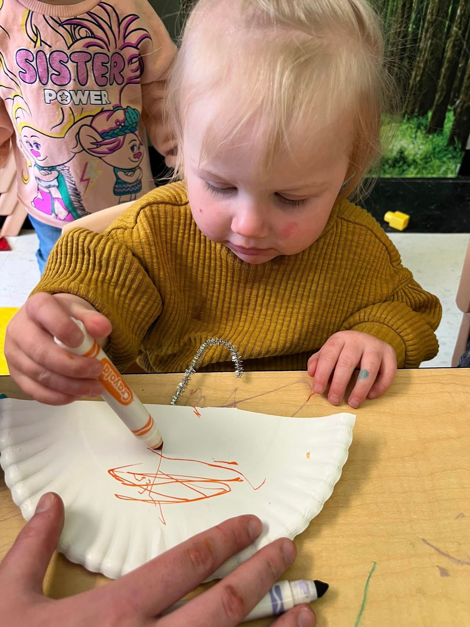 A little girl is sitting at a table drawing on a paper plate with a marker.