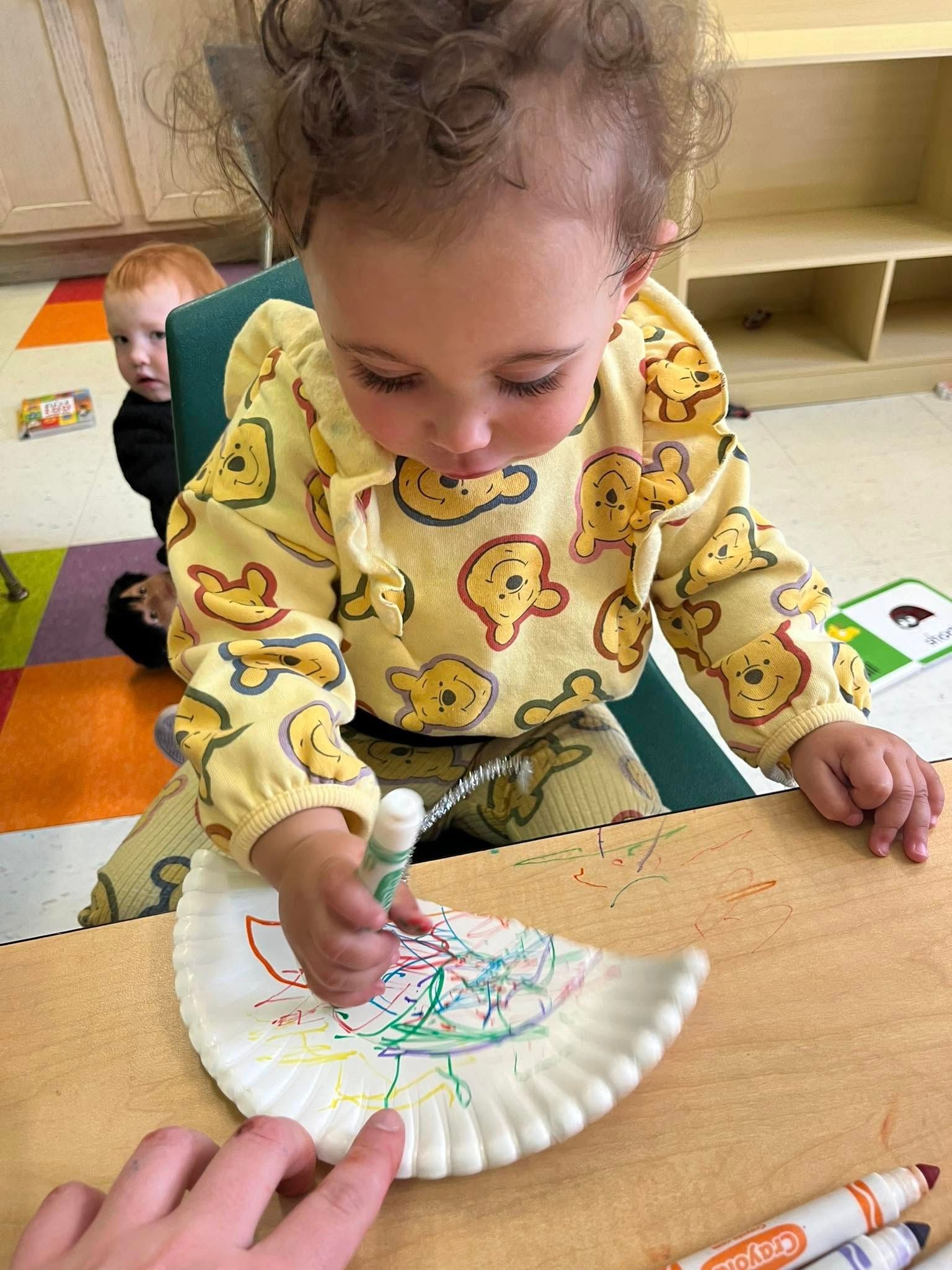 A little girl is sitting at a table drawing on a paper plate.