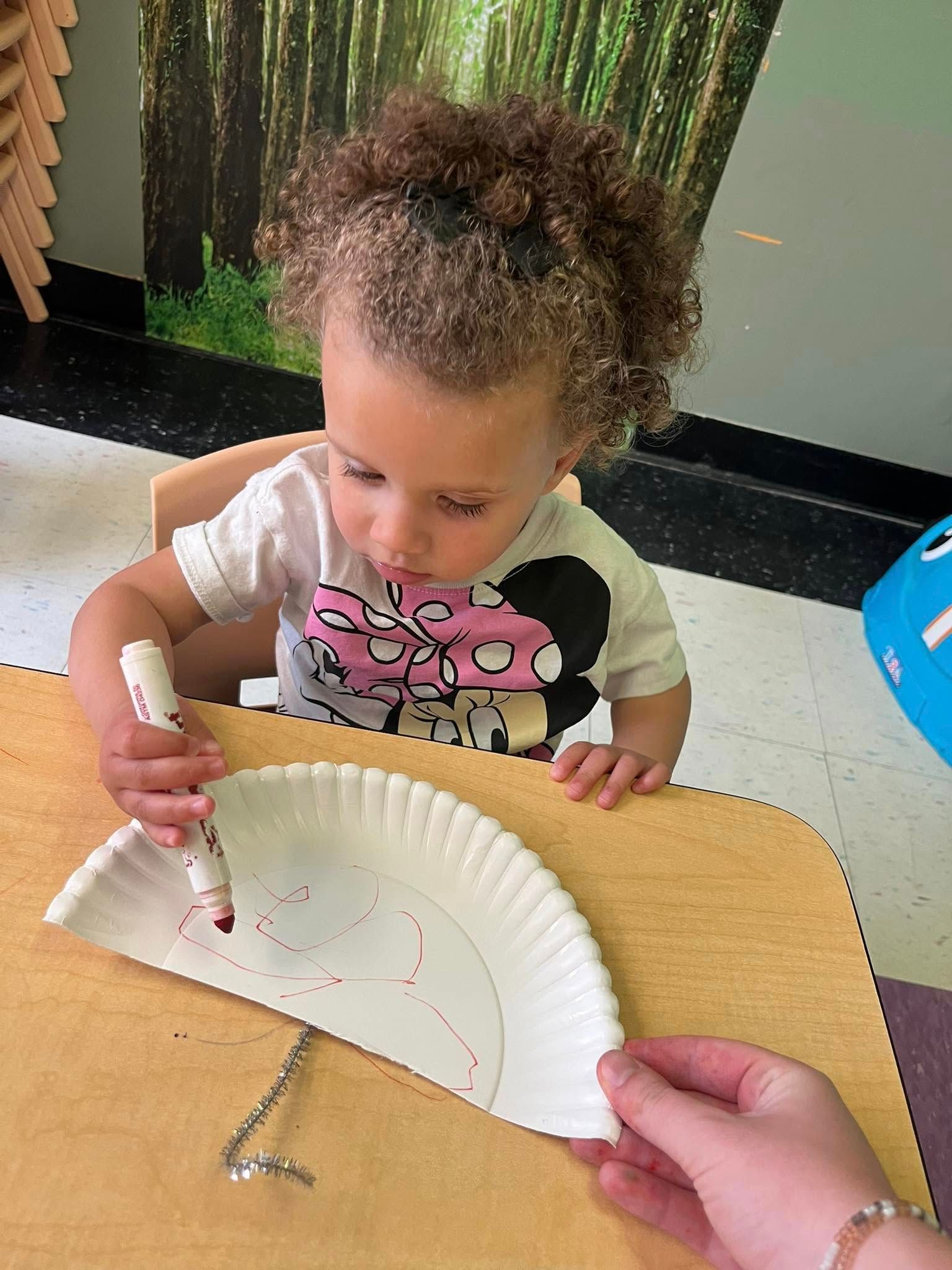 A little girl is sitting at a table drawing on a paper plate with a marker.
