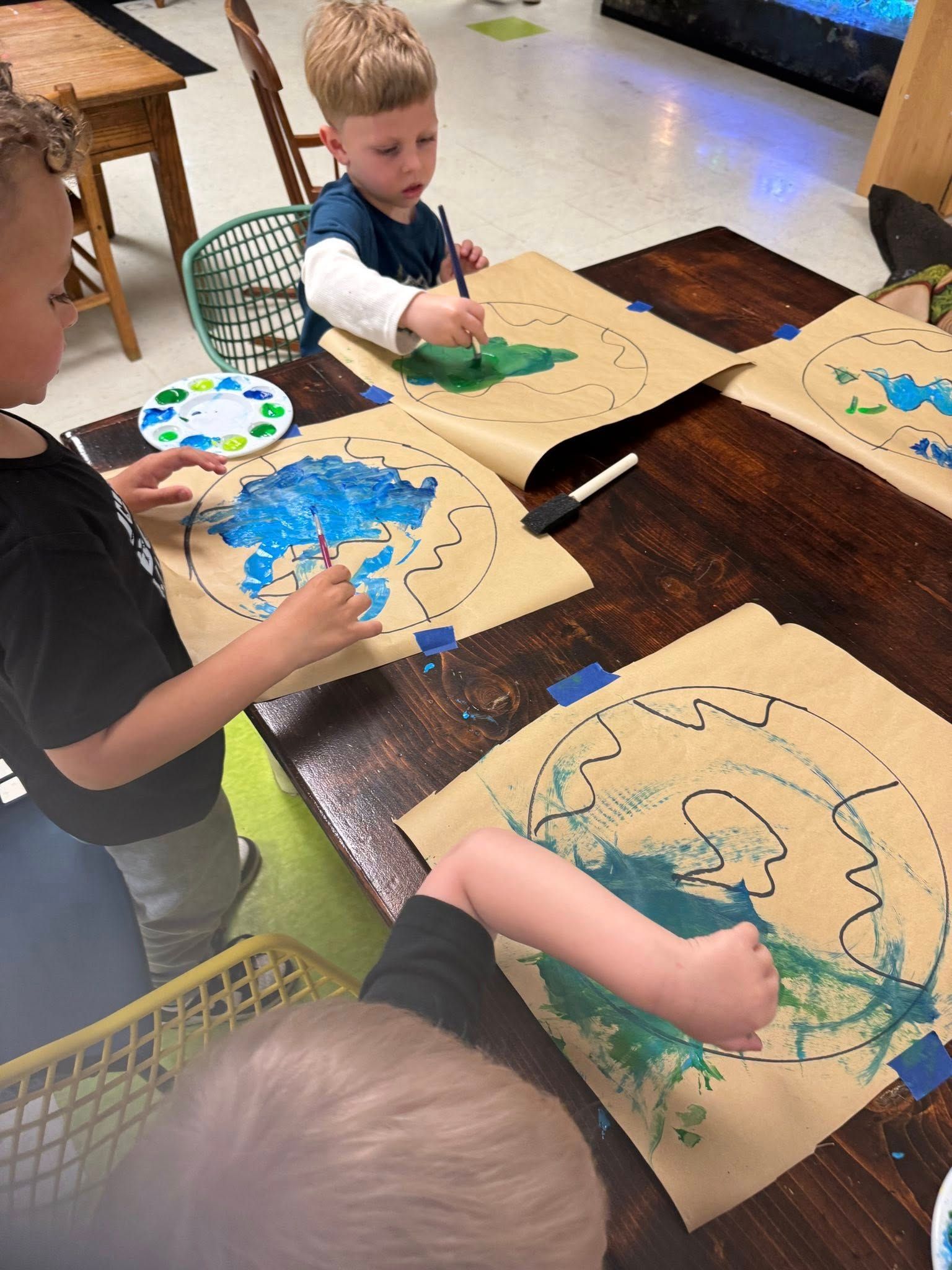 A group of young children are sitting at a table painting on brown paper.