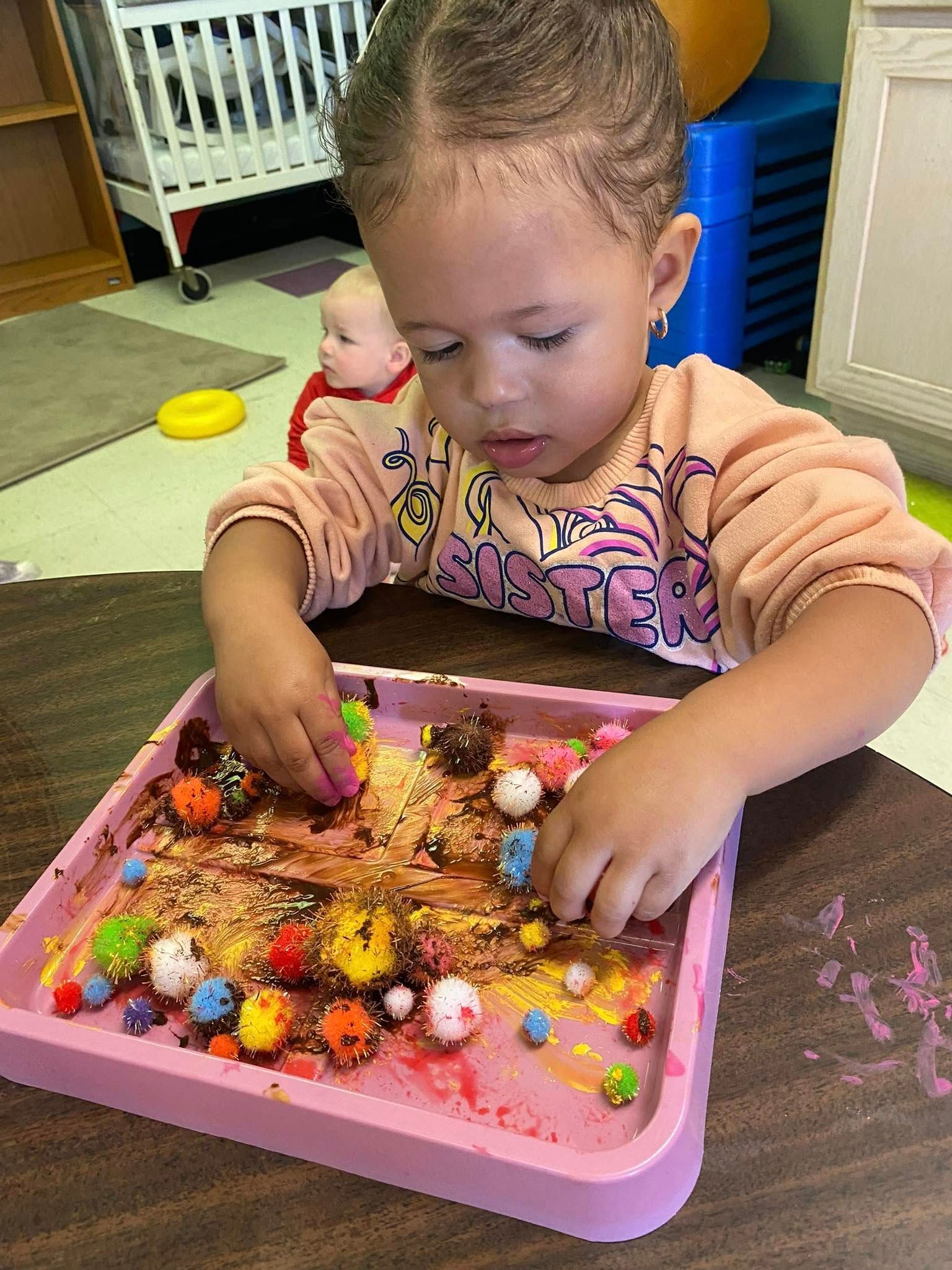 A little girl is sitting at a table playing with a tray of food.